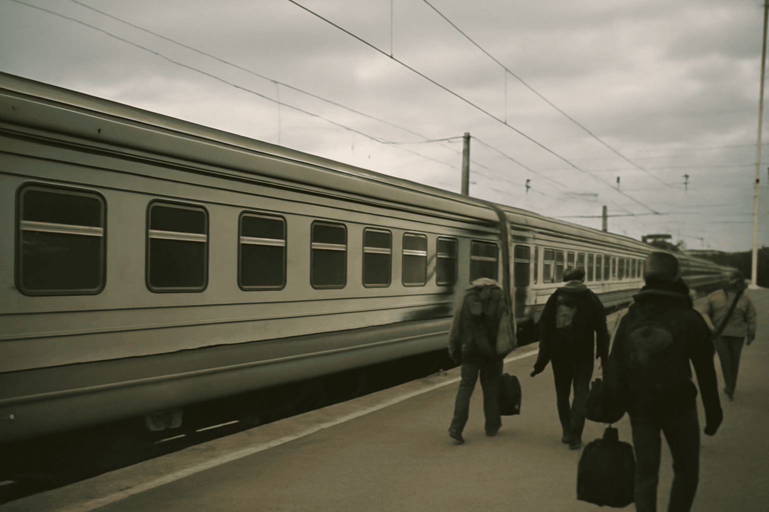 A passenger train waits at a station platform while travellers with bags walk past it under an overcast sky.