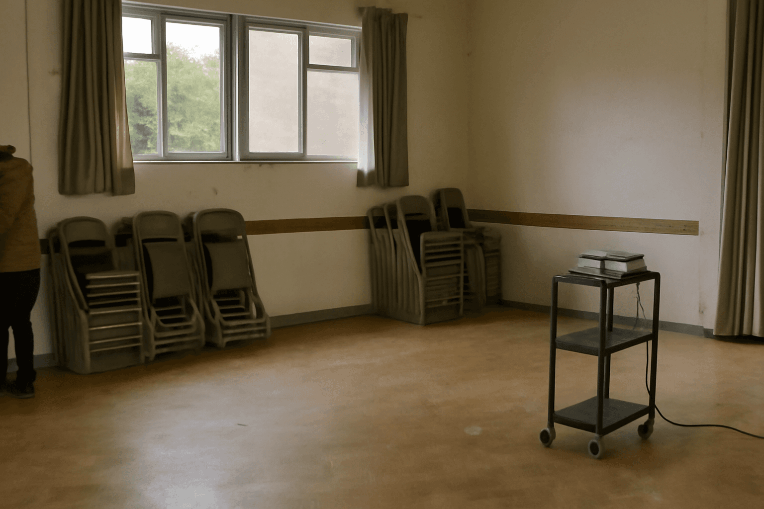 An empty community hall after an event, with folding chairs stacked against the wall and a projector left on a trolley.