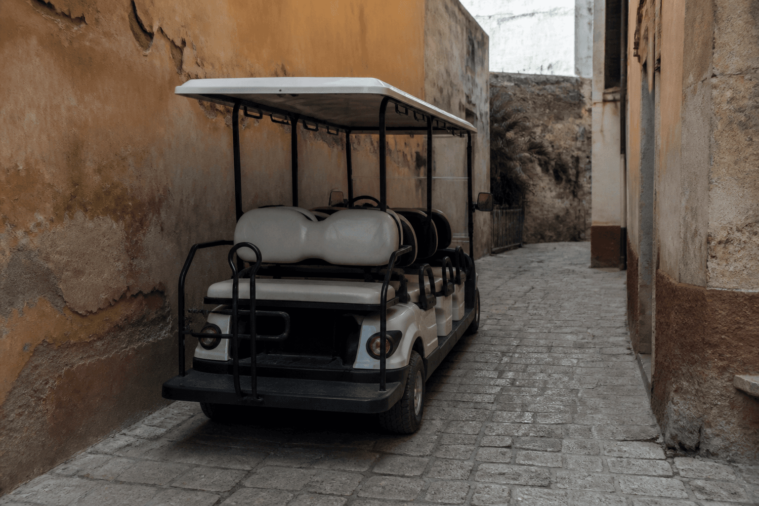 An open-sided golf cart with empty passenger seats parked on a narrow cobblestone street between ochre walls, seen from behind.
