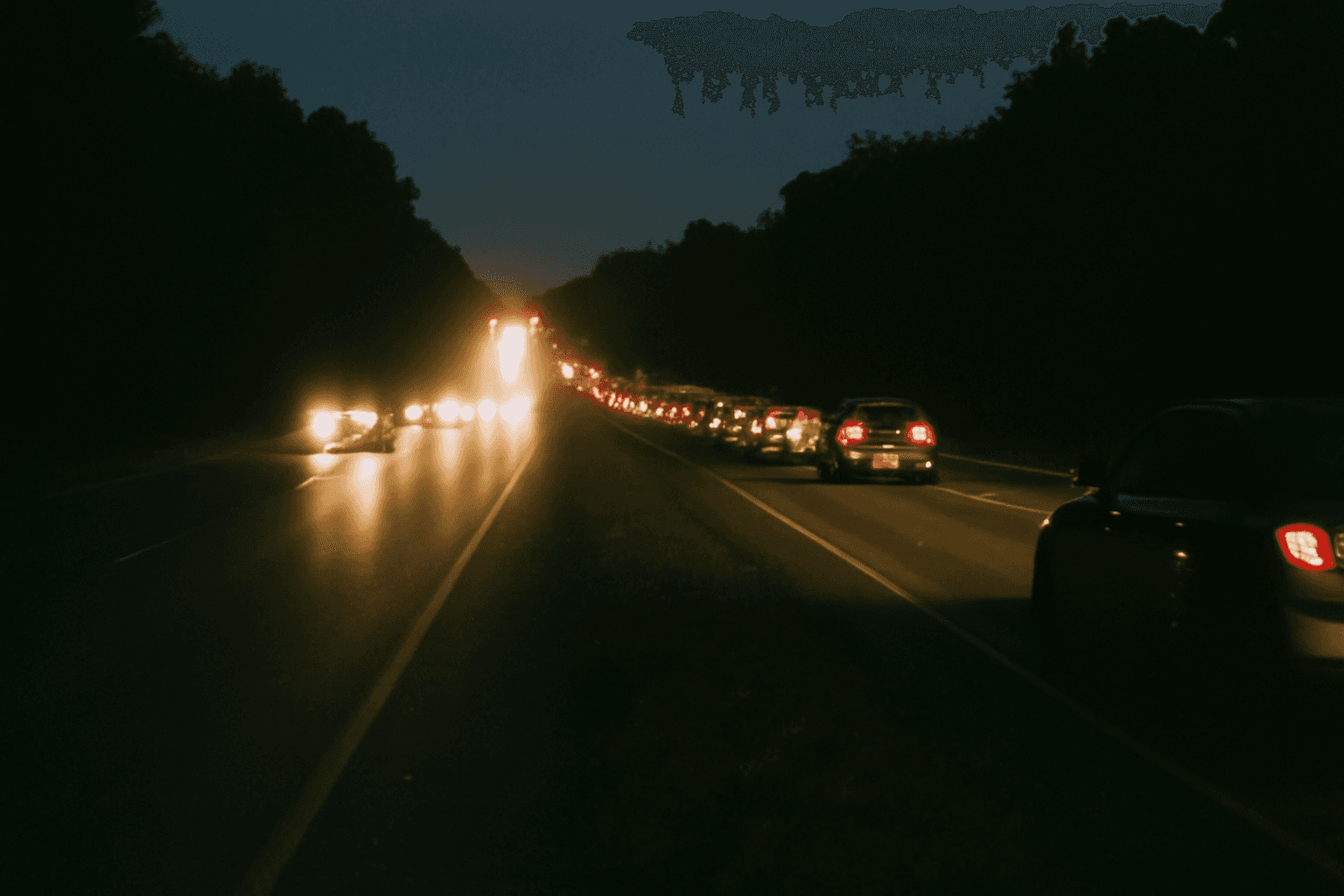 A long line of vehicle headlights streaming down a dark rural highway at night, photographed from the roadside verge.