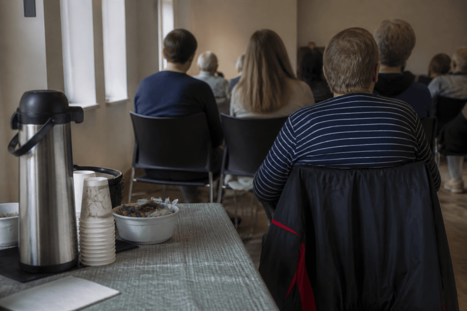 Voters seen from behind seated in rows of chairs in a plain community-hall room, coffee cups on a side table, during a market-research discussion.