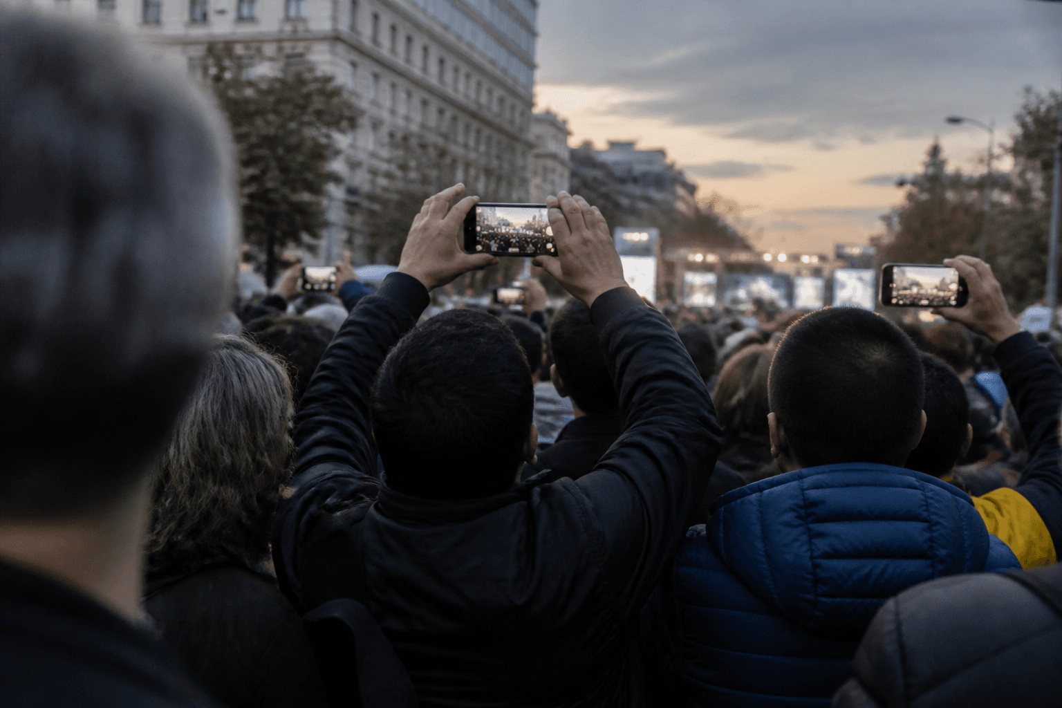 A dense crowd of campaign supporters, seen from behind, fills a city avenue at dusk with phones raised toward a distant stage.