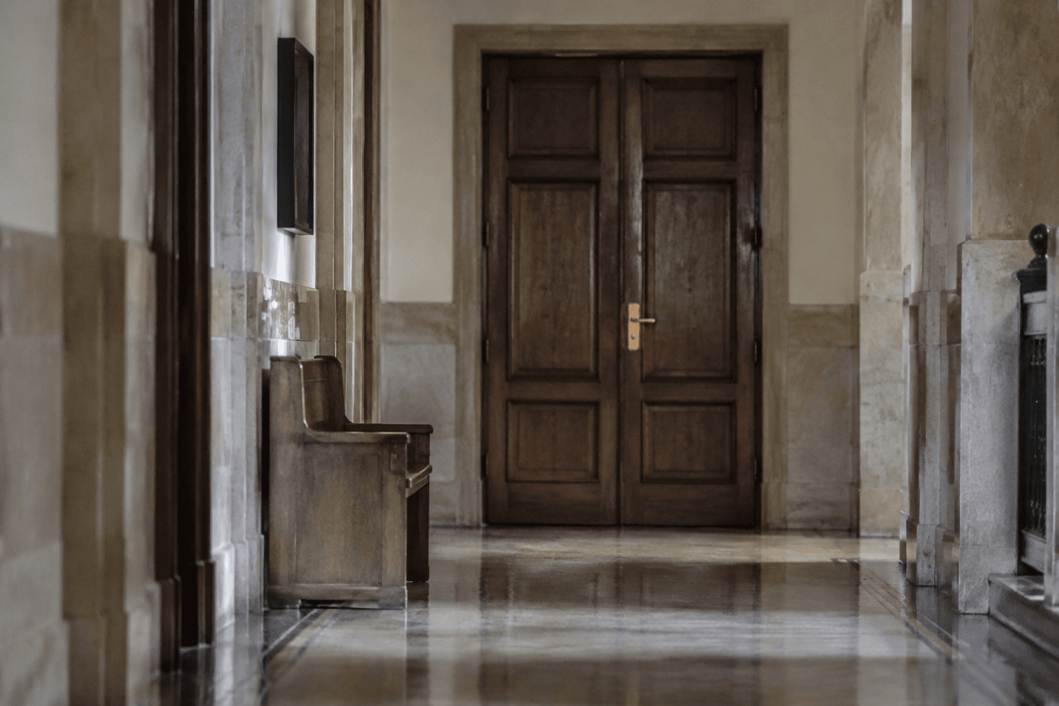 Closed wooden doors with a brass handle at the end of a quiet courthouse corridor, seen empty from a distance.