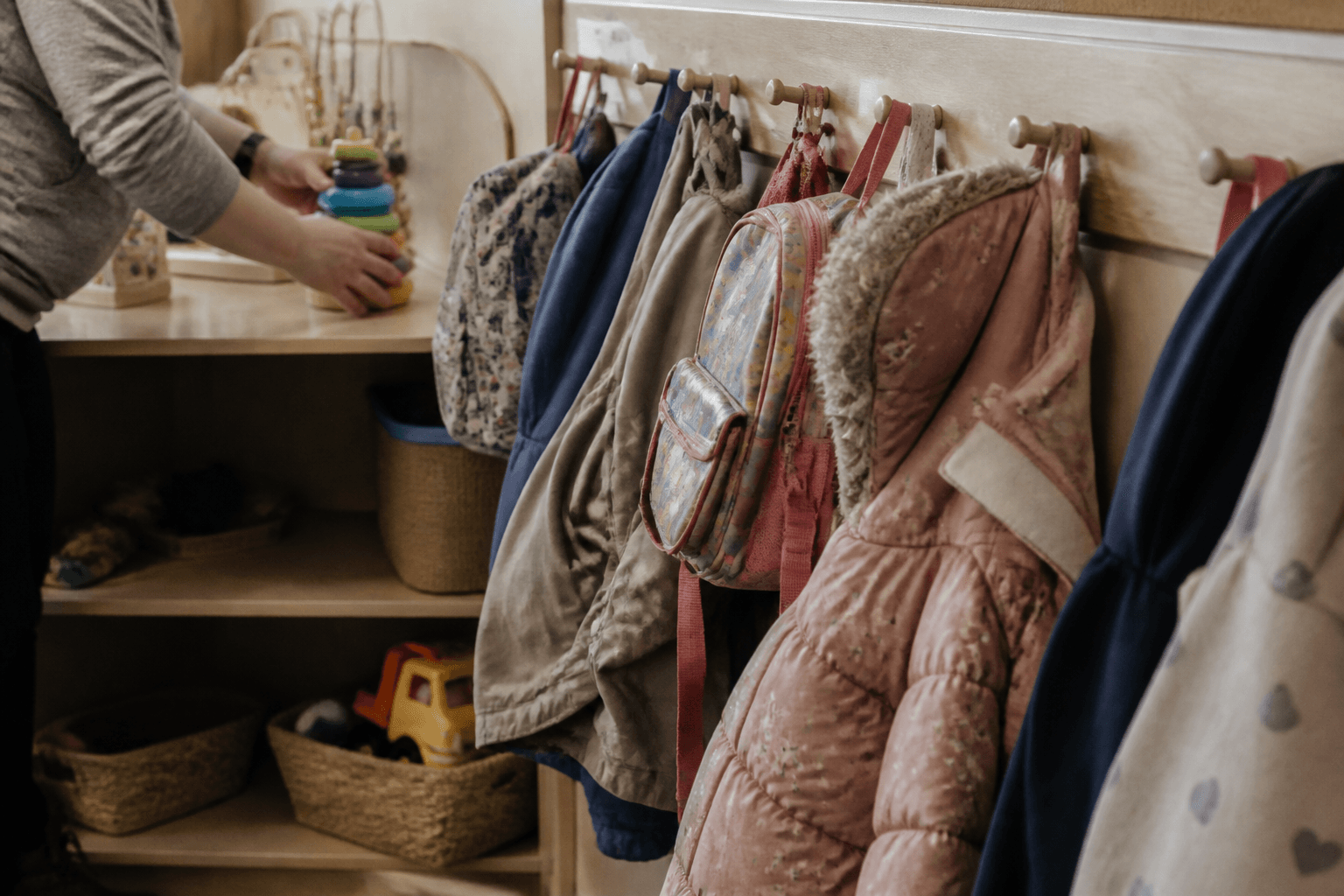 Small coats and backpacks hang from low pegs in a nursery cloakroom while a carer's hands stack toys on the shelf beneath.