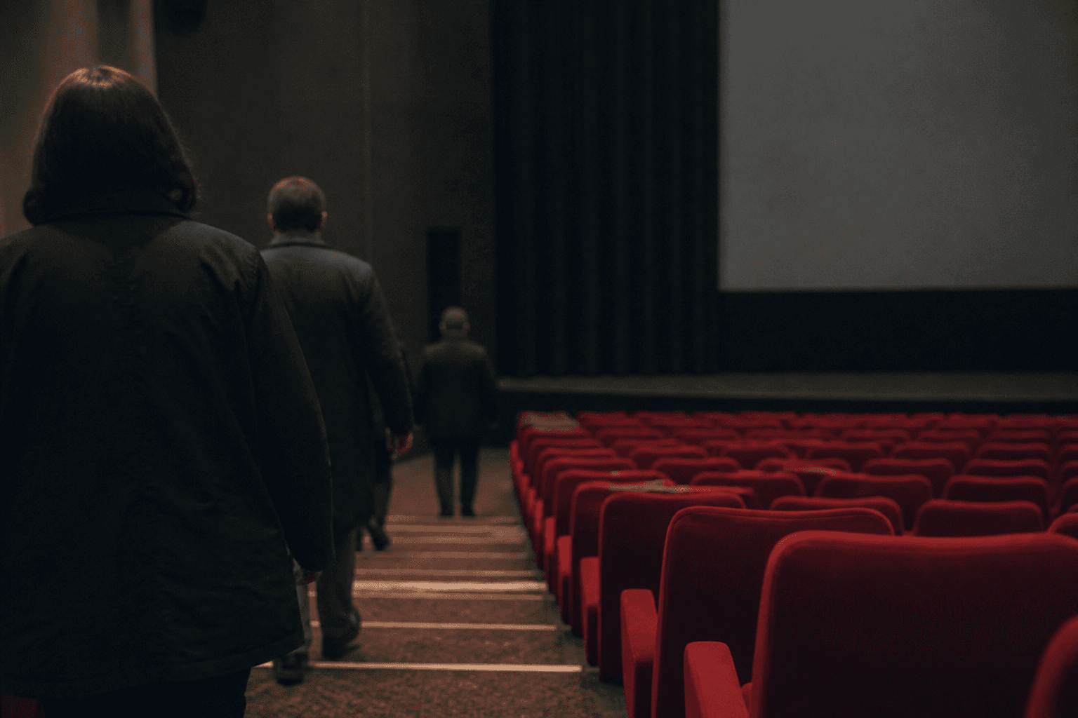 Red cinema seats in a dim auditorium as a few figures seen from behind make their way down the aisle before the film begins.