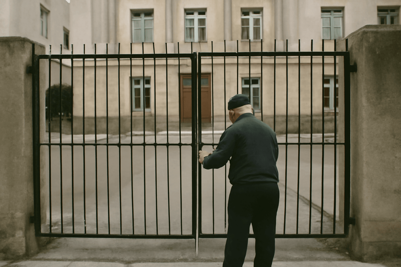A closed metal gate outside a provincial government building on a quiet street, with a security guard seen from behind checking the lock.