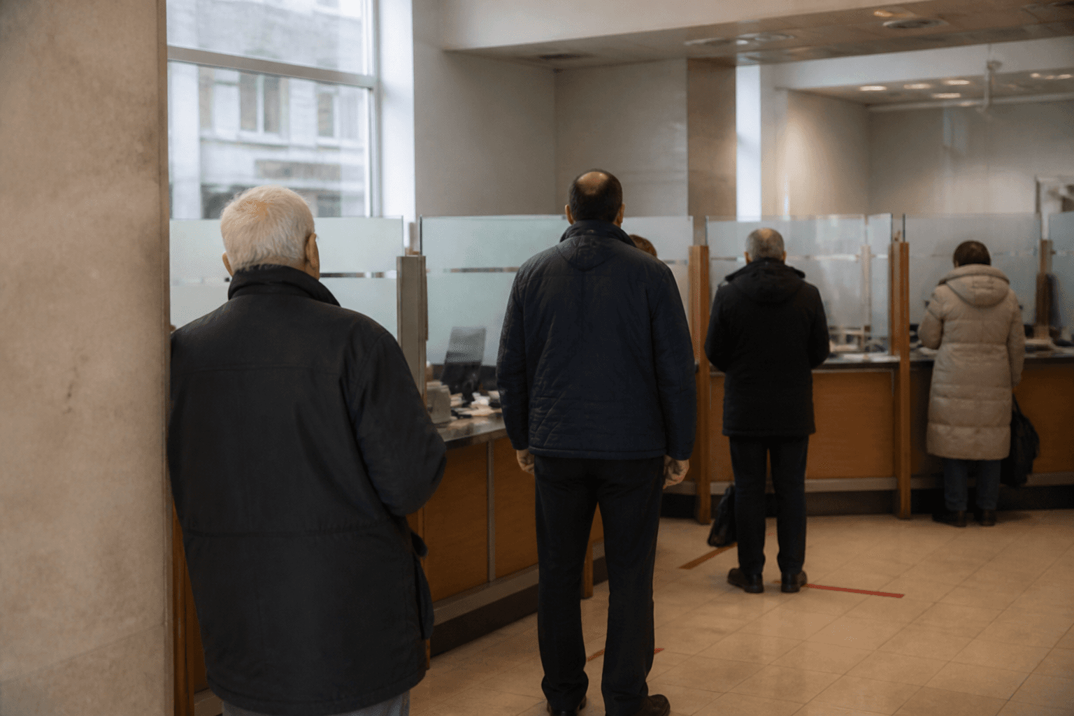 Customers seen from behind wait in a queue at the counter of a high street bank branch on a grey morning.