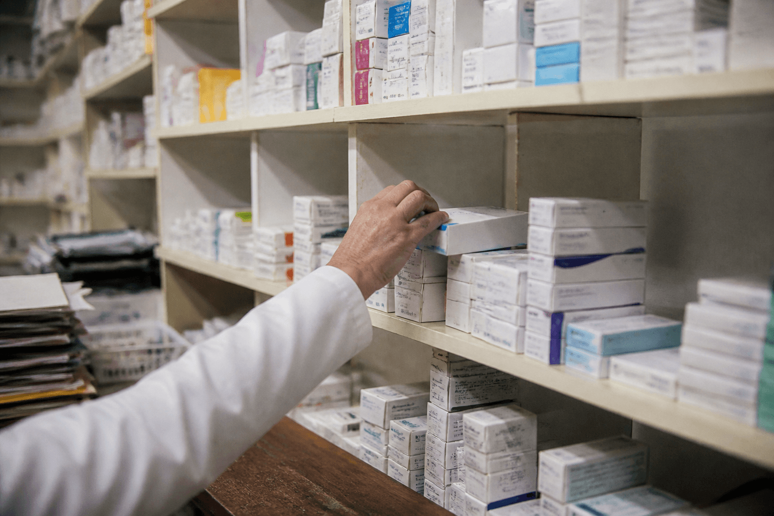 Rows of plain medicine cartons line a shelf behind the counter of a quiet community pharmacy, a hand reaching for one box.