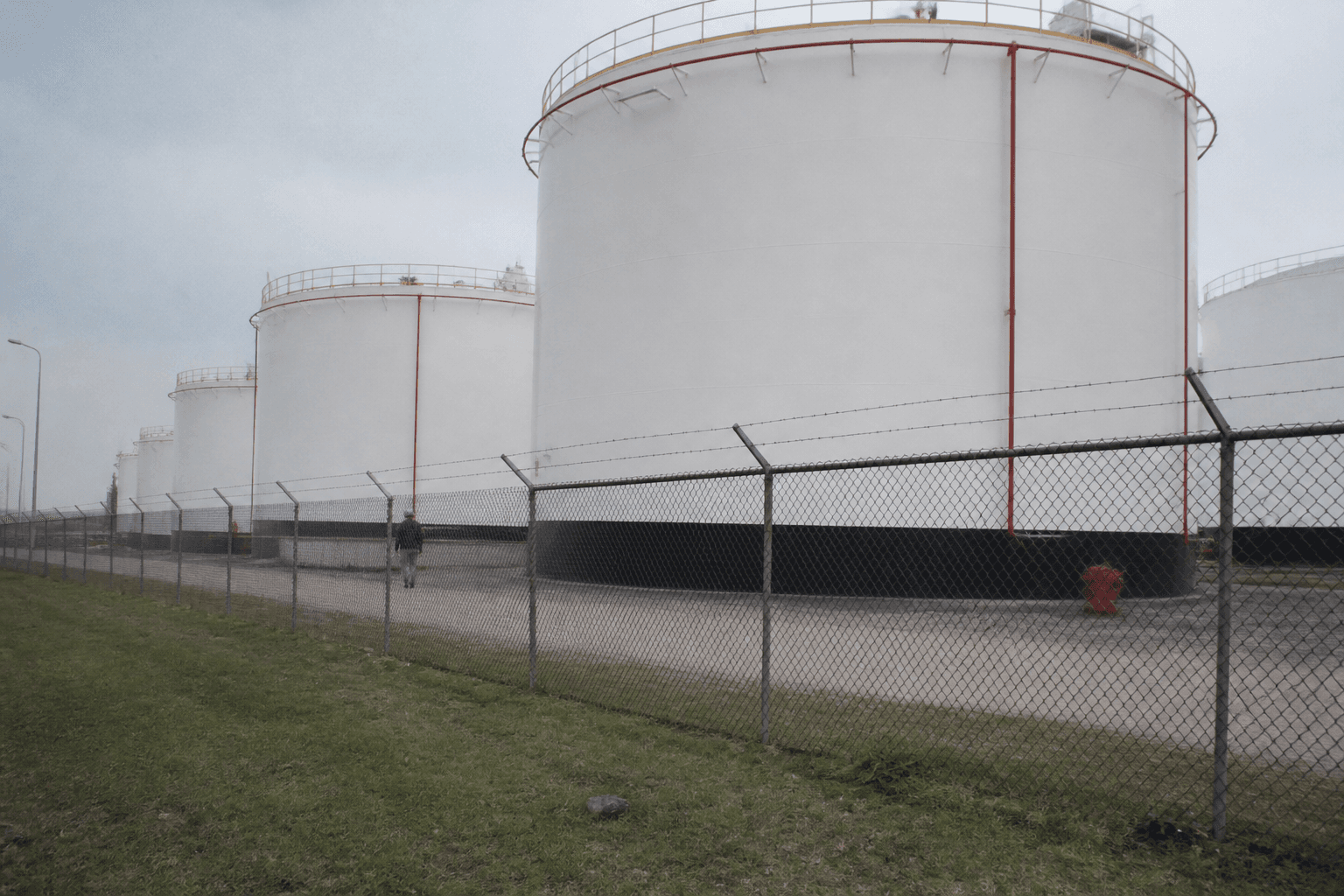Rows of fuel storage tanks behind a chain-link fence at an oil depot, a lone worker seen from far away walking the perimeter road.
