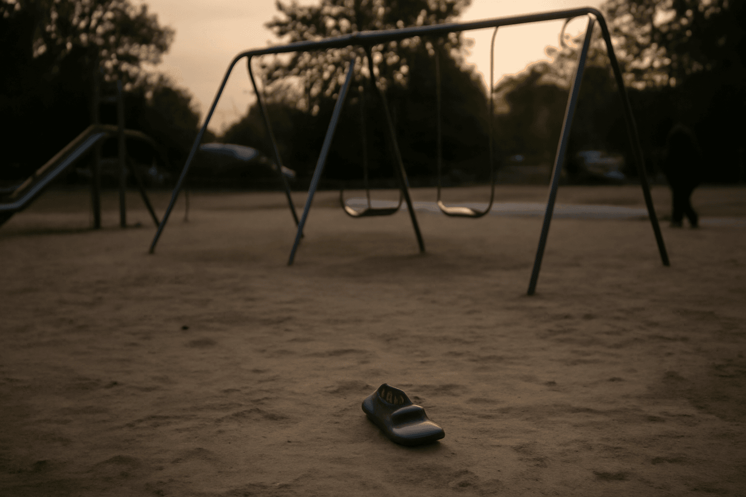 An empty playground at dusk with a small shoe left on the ground near the swings.