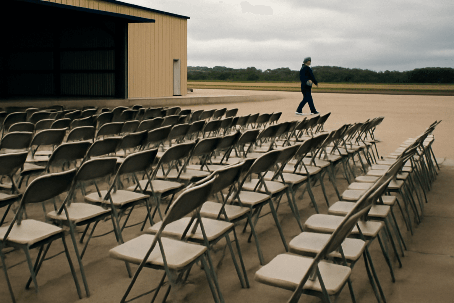 Rows of folding chairs on the tarmac apron of a small regional airport hangar before a political rally, with a figure seen from far away walking past.